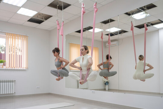 Two women practicing stretching and meditation exercises suspended in pink hammocks during an aerial yoga studio session