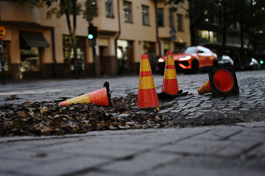 Photograph of traffic safety cones lying on a cobblestone street, with a blurred sports car and urban buildings in the background.