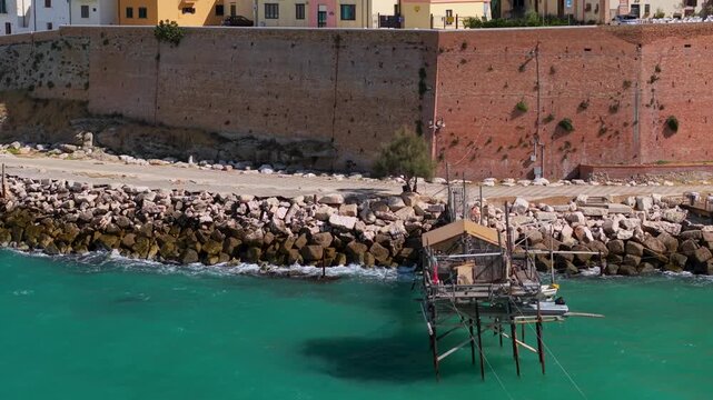 70mm aerial tilt up reveal of Trabucco on water to Termoli Old Town castle walls, Molise Italy over turquoise Adriatic Sea, wooden fishing platform and medieval town B-roll