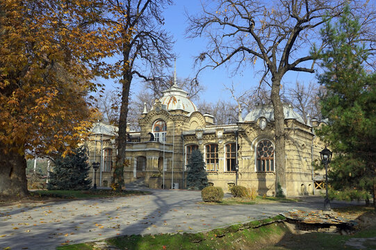 The historic brick facade and ornate spired dome of the Palace of Prince Romanov stand framed by mature autumn trees and a paved courtyard in Tashkent, Uzbekistan