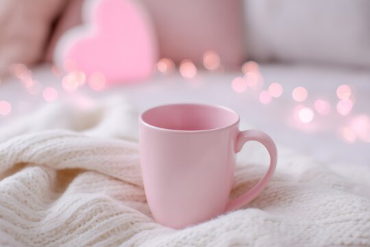 Pastel mug on white knitted blanket with bokeh lights and heart decoration. Romantic Valentine's Day background