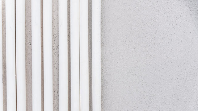 Multiple white vertical plastic pipes run parallel against a textured grey exterior concrete wall