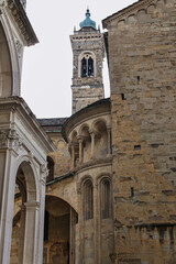Fototapeta premium Historic stone church tower and ornate arched facade seen from narrow courtyard with weathered masonry, blue cupola, classical architectural details, Bergamo