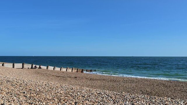 Hayling Island Hampshire England UK. 05.09.2026. Video. People sit peacefully on the shingle shoreline under a clear sky while gentle waves lap against the wooden sea defences on a sunny day