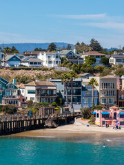 Fototapeta premium Houses and the historic wooden wharf sit beside the sandy shoreline at Capitola Beach in California, where people enjoy water sports and coastal recreation on a sunny day.