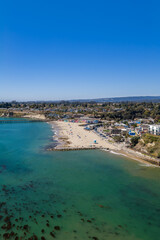 Fototapeta premium Aerial perspective reveals Capitola beach alongside the wooden wharf, beachfront village architecture, shoreline kelp patches, and coastal hills in California under a clear daytime sky.