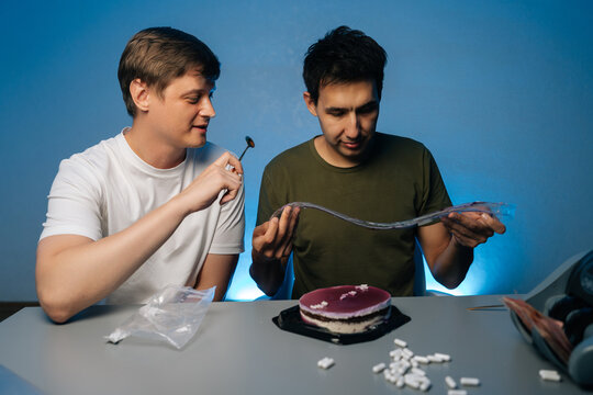 Two young men sitting at table with cake, pills and drug paraphernalia, preparing drugs in home setting, during party with vibrant blue backdrop. Concept of party atmosphere with substance abuse.
