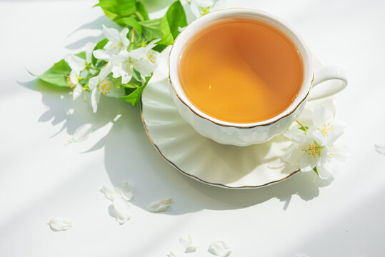 Cup of hot tea with white jasmine flowers and green leaves on a white surface with soft shadows