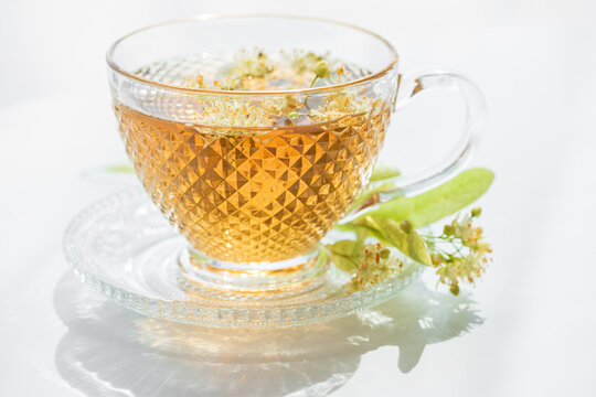 Brewed linden flower tea in glass cup with fresh blossoms and leaves on white background