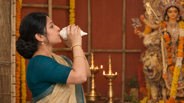 Devout woman blowing conch shell before goddess idol in decorated temple during durga puja navratri and diwali celebrations wearing traditional bengali saree