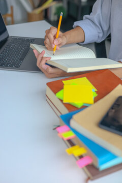 University Student Studying and Writhing in Notebook Preparing for Exam with Textbook on the Table at Home.