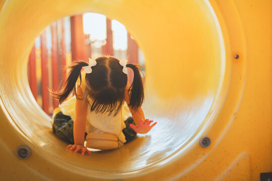 Toddler girl crawling through colorful playground tunnel outdoors, exploring play equipment with curiosity while developing movement, coordination, confidence.