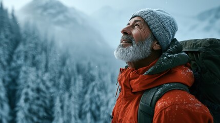 Fototapeta premium Elderly man with gray beard wearing warm winter clothing and backpack enjoying snowy mountain landscape on a cold winter day