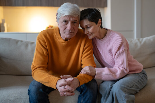 Elderly couple sitting together on couch, compassionate wife embraces worried husband and holds forearm, caring woman gives silent support to distressed partner during difficult life period.