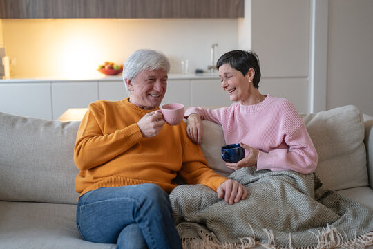 Smiling elderly husband and wife sit close on sofa with warm drinks, affectionate married couple enjoys conversation, mutual attention, companionship and comfortable daily life in retirement.