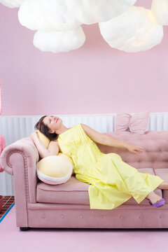 Young woman reclining on pink sofa in pastel living room wearing yellow dress and purple sandals, eyes closed in calm fashion pose