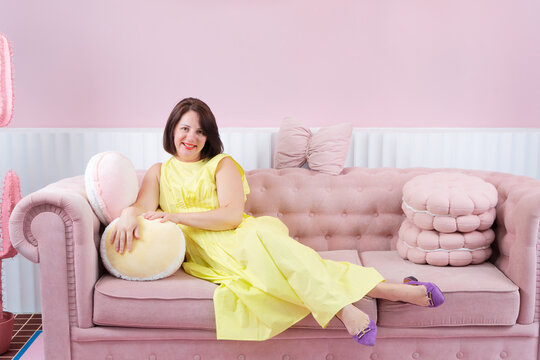 Young woman reclining on pink sofa in pastel living room wearing yellow dress and purple sandals, eyes closed in calm fashion pose
