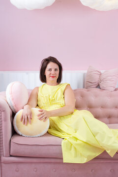 Young woman reclining on pink sofa in pastel living room wearing yellow dress and purple sandals, eyes closed in calm fashion pose