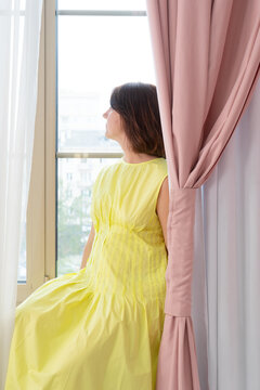 Young woman in white dress sitting on windowsill, gazing outside through sheer curtains in softly lit bedroom, relaxed pose, crossed ankles, delicate interior mood