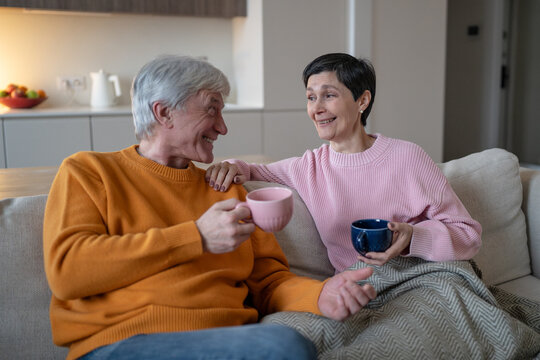 Senior couple sitting on couch with cups, smiling husband and wife look at each other during cheerful conversation, elderly spouses enjoy tea, companionship and quiet retirement leisure.
