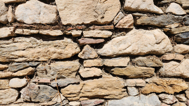 Rough masonry wall built from irregular natural stones of varying sizes, shapes, and colors, with beige, gray, earthy, and terracotta tones, visible mortar joints, and small deposits of soil and veget