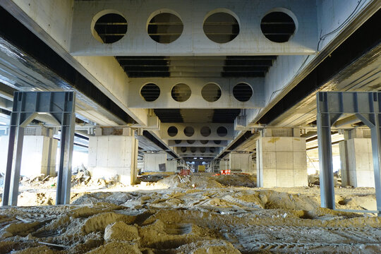 Active construction process ongoing under bridge span. Reveals below-grade building phase, structural repetition, and large-scale civil engineering process on urban transport infrastructure.