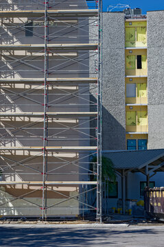 Scaffolding wraps a multi-story apartment building undergoing exterior renovation, showing yellow insulation panels, metal platforms and ladders against a bright blue sky, suggesting construction, rep
