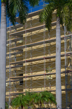 Scaffolding wraps a multi-story apartment building undergoing exterior renovation, showing yellow insulation panels, metal platforms and ladders against a bright blue sky, suggesting construction, rep