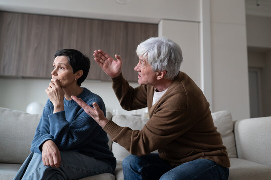 Irritated husband aggressively arguing with emotionally distanced elderly wife in living room, senior man in despair angry gesturing and shouting at exhausted woman, domestic argument, family conflict
