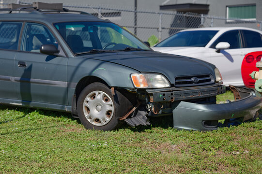 Close-up of a damaged car front bumper showing rusted metal frame, broken headlight, detached fender and scratched paint, conveying vehicle damage, repair needs and roadside wear and tear.
