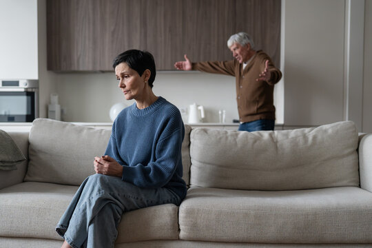 Senior marriage crisis and relationship problem, elderly angry husband blaming and arguing with wife, stressed woman sitting on sofa while irritated man shouting behind, emotional distance and divorce