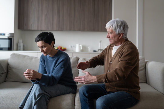 Senior couple in serious argument and misunderstanding sitting together in living room on sofa, angry elderly man speaking to emotionally exhausted woman, relationships problem, family crisis.