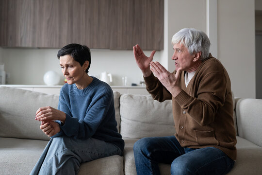 Hysterical aggressive elderly husband arguing and accusing sad depressed wife during quarrel, senior married couple having argument while sitting on sofa at home, aged man yelling at unhappy woman.