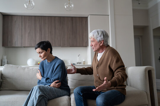 Disappointed frustrated senior husband family quarrel with unhappy sad wife sitting on sofa, disagreement and communication barrier, elderly defensive woman ignoring nonstop accusations from partner.