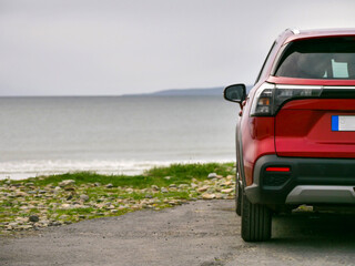 Red SUV parked on coastal gravel path. Ocean waves crash nearby, distant hills visible. Vehicle facing away, rear view prominent. Calm, adventurous vibe for travel or road trip themes © mark_gusev