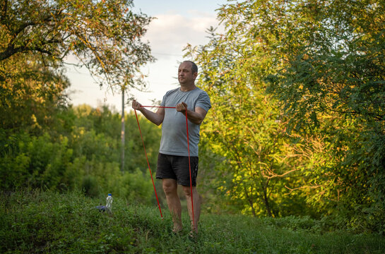 Barefoot man trains with red exercise band in wooded greenery, holding tension with both hands and determined expression. Outdoor resistance workout takes place on quiet grassy path.