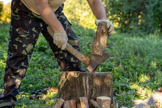 Man steadies log while splitting firewood with axe on tree stump in sunlit woodland. Camouflage trousers and protective gloves suit outdoor wood chopping among grass and leafy plants.