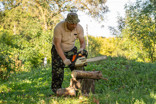 Man holds chainsaw over logs in woodland, focused on cutting firewood. Wearing camouflage trousers and bucket hat, worker steadies power tool beside tree stump in sunlit green clearing.