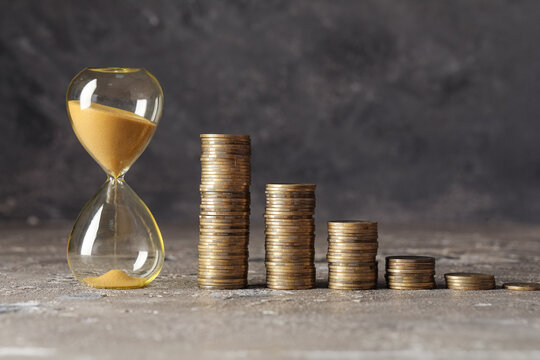 Time is money concept, hourglass with falling sand next to decreasing stacks of gold coins financial planning and investment deadline