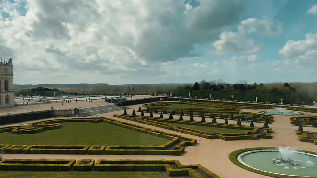 Aerial view of a magnificent French formal garden featuring manicured green parterres, decorative fountains, symmetrical hedges, and an imposing historic royal palace under a scenic cloudy blue sky.

