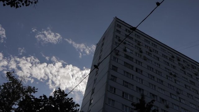 An urban landscape. An old gray panel building and clouds drifting across a blue sky in the background; in the foreground, a pigeon lands on power lines. Silhouettes of birds on a sunny day