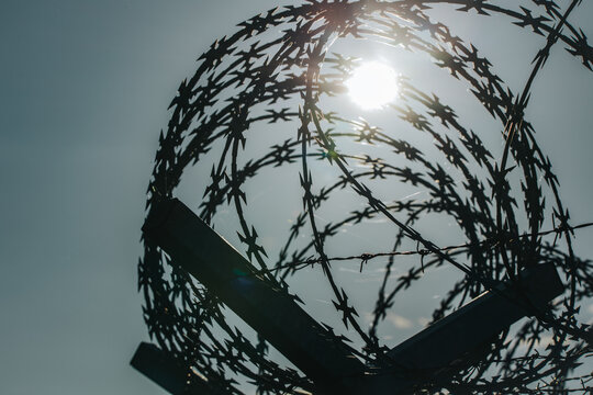 Barbed wire coils silhouetted against bright sun in blue sky with angular support beams, creating a stark abstract industrial security scene