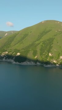 Drone footage of Kezenoyam Lake surrounded by green mountain slopes and rocky shores in the Caucasus Mountains, Chechnya Republic, Russia, on a clear day