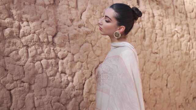 Cinematic portrait of an elegant woman in a patterned shawl touching a textured mud wall in AlUla