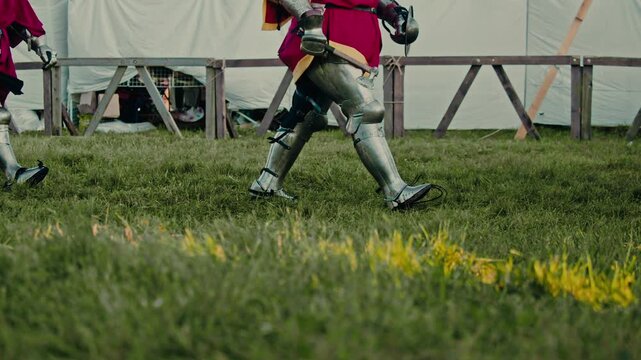 Armored knights walk on grass carrying sword and axe. Brave warriors wear red surcoats over heavy metal leg protection while marching toward brutal medieval tournament battle.