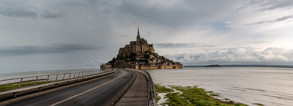 Road to Mont-Saint-Michel during high tide with rising water, tidal phenomenon on Atlantic's ocean coast, view on UNESCO heritage site and iconic Normandy landmark and green pastures