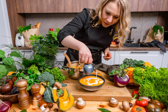 Autumn lifestyle cooking &mdash; woman with pumpkin comfort soup. Culinary enjoyment and homemade nutrition concept. Focused female cook in apron pouring fresh cream from jug into bowl with yellow puree.