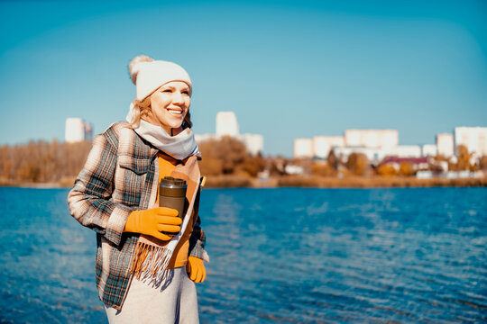 Relaxed woman holding coffee in autumn park near river &mdash; perfect city escape. Happy traveler enjoying weekend nature walk. Calm outdoor recreation restoring mental health balance.