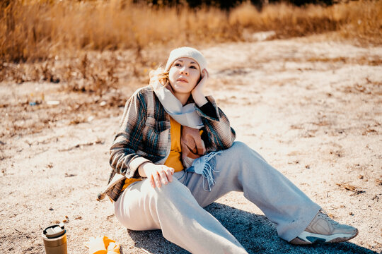 Thoughtful woman resting on autumn river coast &mdash; profound mental health recovery. Calm traveler sitting on sand enjoying long park walk. Peaceful outdoor hiking journey far from urban life.