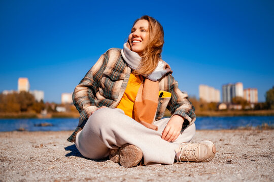 Cheerful woman enjoying bright autumn sunlight in park &mdash; positive wellbeing concept. Happy hiker resting on river sand. Wonderful outdoor walk providing perfect mental health escape.